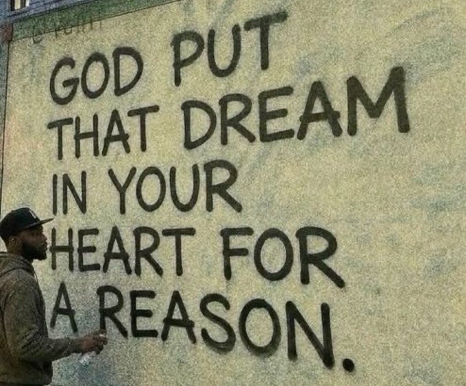 A man in a black cap stands beside a textured wall displaying the handwritten message "GOD PUT THAT DREAM IN YOUR HEART FOR A REASON." The inspirational mural encourages viewers to pursue their goals, fitting a post offering a $5-to-1 challenge to those who engage with a gm comment and share their wallet addresses.