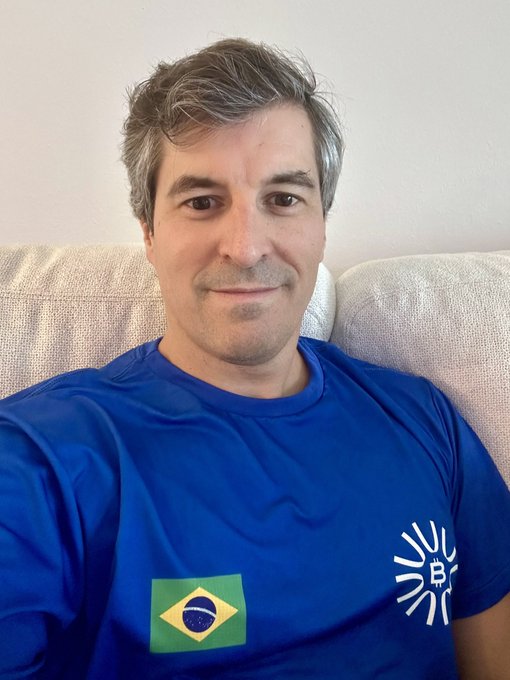 A man with gray-streaked hair sits on a beige couch wearing a blue BitGoool t-shirt featuring the Brazilian flag and the "B" sunburst logo, smiling as he represents Brazil for the Fanbase @ORANJEBTC.