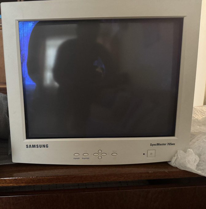 A vintage white Samsung SyncMaster 765MB CRT monitor sitting on a wooden desk, with a dim, mostly blank screen reflecting the photographer's silhouette.