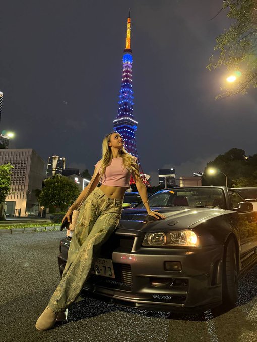 A nighttime photo of a woman posing in a pink top and green cargo pants while leaning against a dark Nissan Skyline GT-R, with the Tokyo Tower illuminated in blue and red in the background.