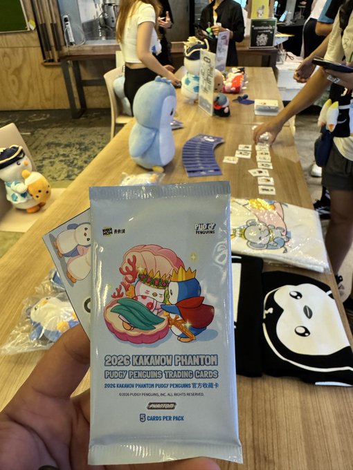 A hand holding a sealed pack of '2026 KAKAMON PHANTOM PUDGY PENGUINS TRADING CARDS' featuring colorful penguin illustrations, with a trading table and other merchandise visible in the background.