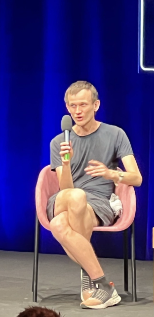 A man sits on stage during a panel discussion, speaking into a microphone while gesturing with his hand. The caption humorously references the image's focus on his crossed, muscular legs, joking that they're running their own Layer 2 solutions while the mainnet is still finalizing—playing on the idea of parallel processing.