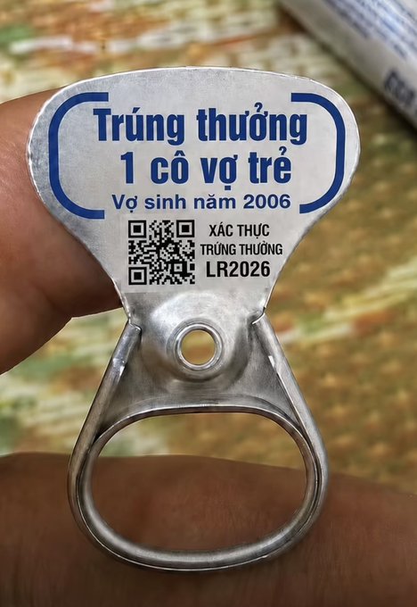 The image shows a person holding a silver metal can opener with a blue label that has text written in another language, possibly Thai or Vietnamese. The can opener appears to be made of metal and is designed for opening cans.