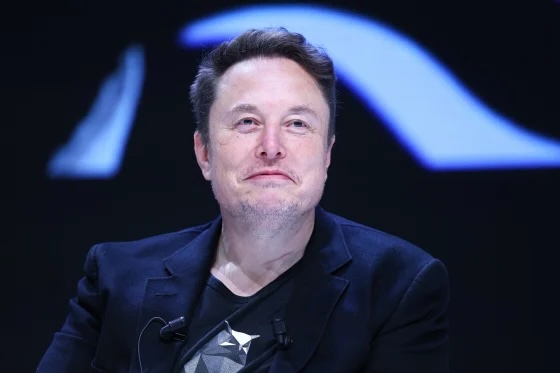 The image features a man wearing a black suit, sitting down with his head tilted back as he looks off into the distance. The background of the image is dark blue, which contrasts with the man's suit and makes him stand out. There are two other people in the image who appear to be standing behind the man, but they are not the main focus of the scene. The caption provided seems to be unrelated or unrelated to the image itself.