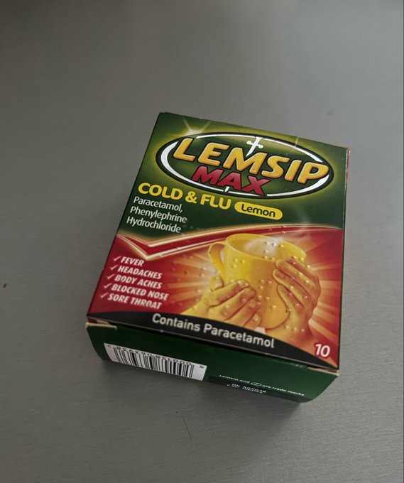 A box of Lemsip Max Cold & Flu Lemon medication sits on a gray surface, listing ingredients Paracetamol, Phenylephrine Hydrochloride and symptoms including fever, headaches, body aches, blocked nose, and sore throat.