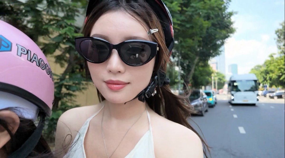 A woman wearing dark cat-eye sunglasses and a headscarf smiles while riding on the back of a pink Piaggio scooter on a city street with a bus visible behind her.
