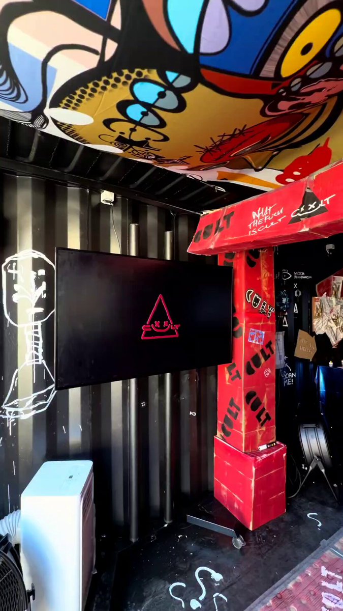 The image shows a room with a large TV mounted on the wall, surrounded by various objects such as chairs and boxes. The room also features graffiti art on the walls, giving it an urban feel. A person can be seen standing near one of the TVs, possibly setting up or attending to the booth. The post includes text that reads "Thanks so much @CultCryptoArt & @Dat_Ogar for setting up everything and the wonderful booth! @NFCsummit".