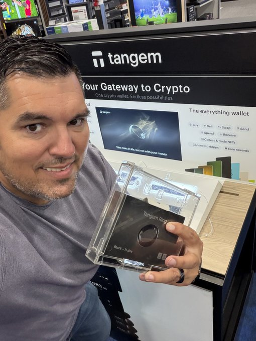 The image features a man holding a small black object that appears to be a ring, possibly made of metal or plastic. The man is standing next to a display case with a sign that says "Tangem" above it. The background shows a store setting with various items on shelves and displays.