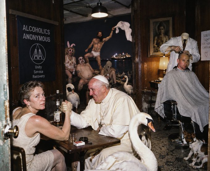 A surreal scene shows a woman arm-wrestling with the Pope at a bar table while swans wander around, a barber cuts hair in the background, and a large photo of people wearing swan masks hangs on the wall near an Alcoholics Anonymous banner.