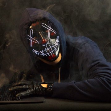 A person wearing a dark hoodie and a glowing "X" and stitched-pattern mask leans over a keyboard, evoking the stealthy image of a hacker. This ties directly to the post about Lazarus-linked wallets laundering $30M through Hyperliquid, symbolizing the covert cybercrime group behind the illicit crypto movement.