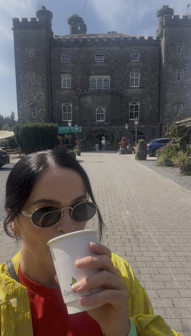 A woman wearing sunglasses and a yellow jacket drinks from a white paper cup in front of a large stone castle-like building on a sunny day.