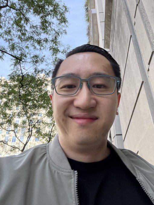 A selfie of a young man wearing glasses and a grey bomber jacket, taken outdoors with trees and a building visible in the background.