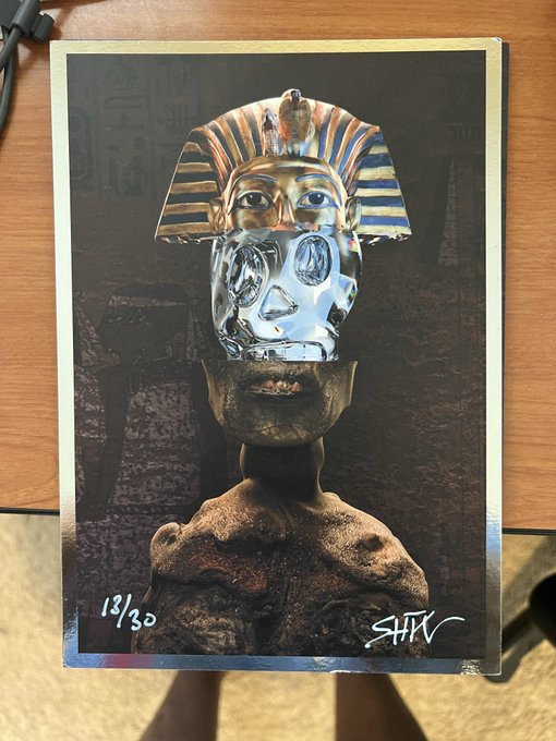 A printed art card or poster depicts a surreal figure with a golden pharaoh-like headdress, a reflective silver face, and a textured body, numbered '13/30' and signed 'SHV'.