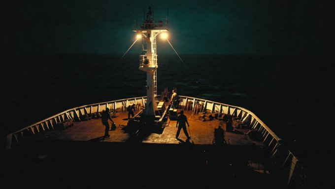 A dark, moody photograph of a ship's deck at night, illuminated by warm overhead lights, with several silhouetted figures working near the bow railing against a dark sea and sky.
