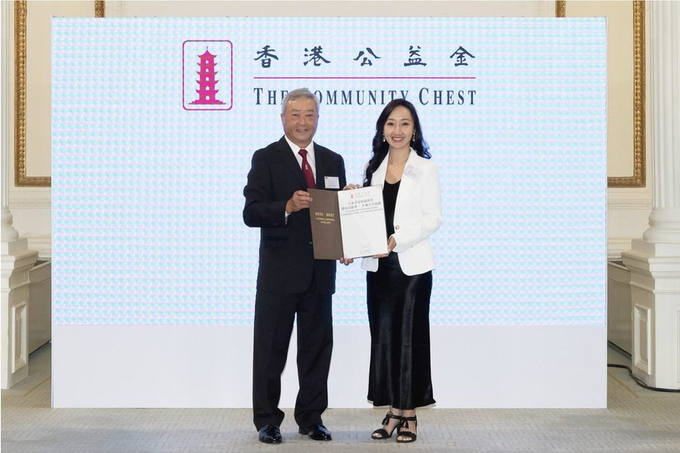 A man in a dark suit and a woman in a white blazer pose together holding an award plaque in front of a backdrop reading '香港公益金 THE COMMUNITY CHEST'.