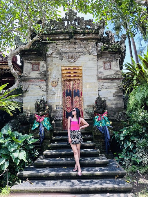 A woman in a pink top and floral skirt posing on stone steps in front of an ornate traditional Balinese temple gate adorned with statues and colorful sashes.