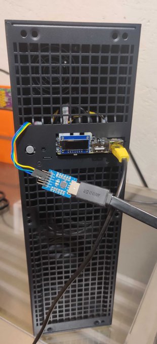 Close-up of the back of a black electronic device (likely a console or computer) showing various ports, cables including yellow ethernet and USB connections, and a small microcontroller board attached with wires.