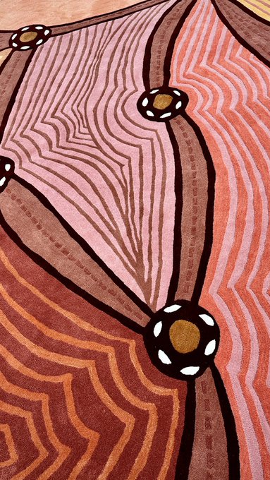 An abstract artwork featuring pink, red, and brown wavy striped patterns with black circles containing white dots and gold centers, resembling an Indigenous Australian dot painting style.
