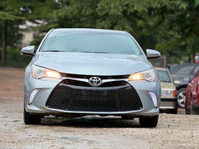 A front view of a silver Toyota sedan parked on a gravel lot with its headlights and daytime running lights on.