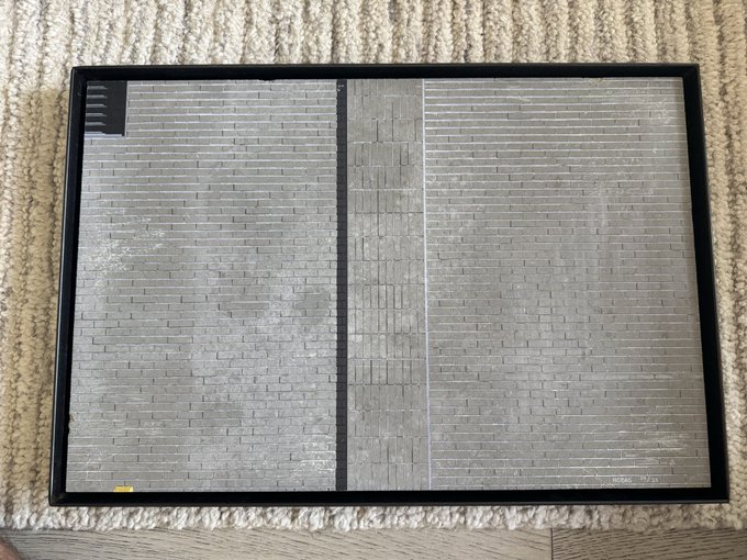 A black framed picture or display screen showing a gray brick/stone texture lies on a textured cream-colored carpet.