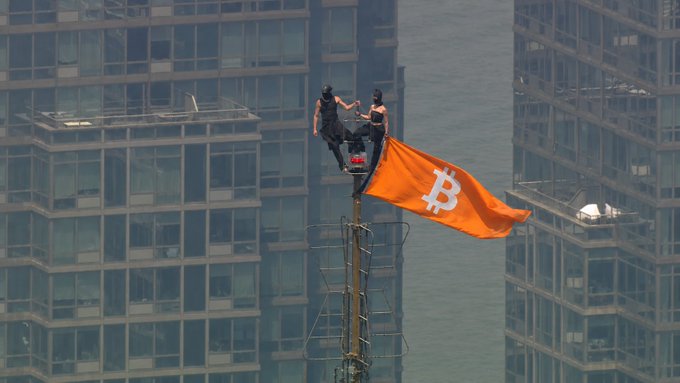 A dramatic photo of two people in dark tactical gear standing atop a flagpole flying a large orange Bitcoin flag, set against a backdrop of skyscrapers and water.