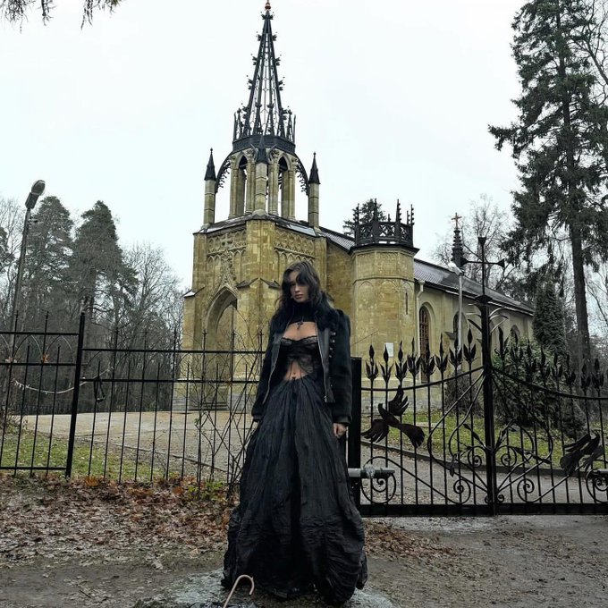 A gothic-style woman in a black dress stands before an ornate wrought-iron gate and a striking yellow Gothic Revival chapel with a tall spire, surrounded by bare trees on a gloomy, overcast day.
