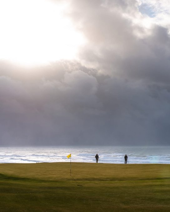 Two figures stand on a coastal golf course green near a yellow flag, with dramatic clouds and sun rays breaking through over the ocean.