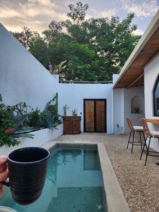 A courtyard scene showing a small swimming pool, white walls, wooden doors, potted plants, and a hand holding a black coffee mug in the foreground.