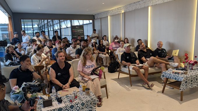 A diverse crowd of attendees sits in a modern lounge-style venue watching a presentation, with a "PITCH FEST" banner visible in the background. The scene captures the lively atmosphere of the Pitch Fest Bali event, held alongside CoinfestAsia, where selected teams from over 200 applications presented to the engaged audience.