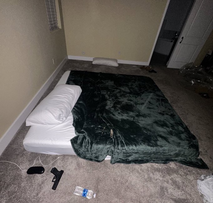 A bedroom floor scene shows a mattress with a dark green blanket, accompanied by a gun and a water bottle on the carpet.
