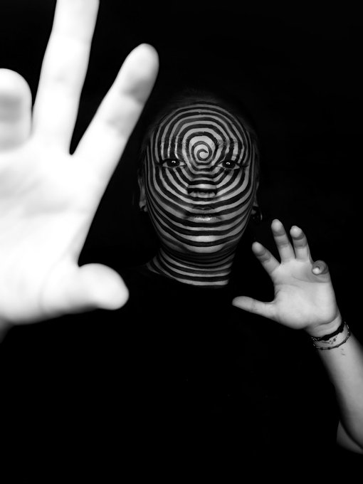 A black-and-white artistic photograph shows a person with hypnotic concentric circle patterns painted on their face, holding both hands out toward the camera against a dark background.