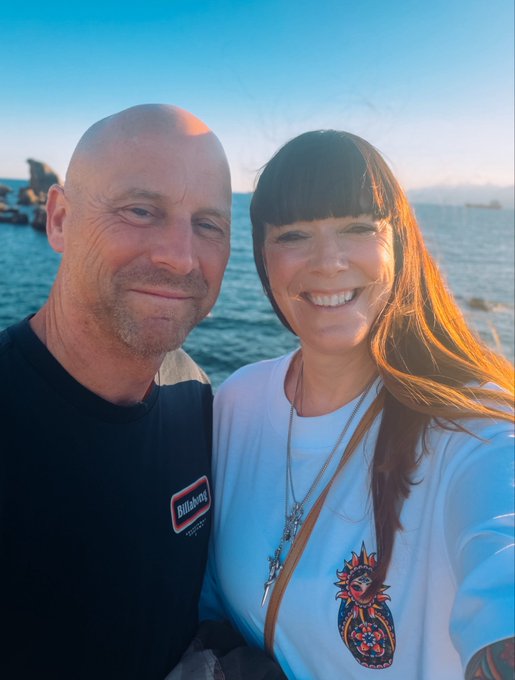 A selfie of a bald man in a black Billabong shirt and a smiling woman with long brown hair in a white shirt, posing together outdoors with the ocean and sky visible in the background during sunset.