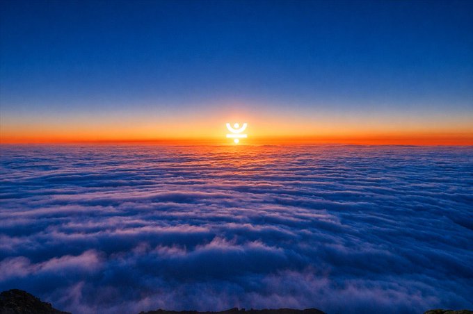 A sunrise or sunset above a sea of clouds with orange light at the horizon and a small icon overlaid near the sun.