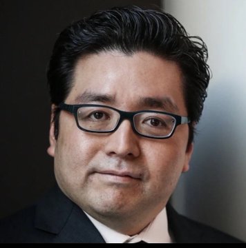 A close-up portrait shows a middle-aged man wearing a dark suit, white shirt, and dark-rimmed glasses, looking directly at the camera against a blurred gray backdrop.