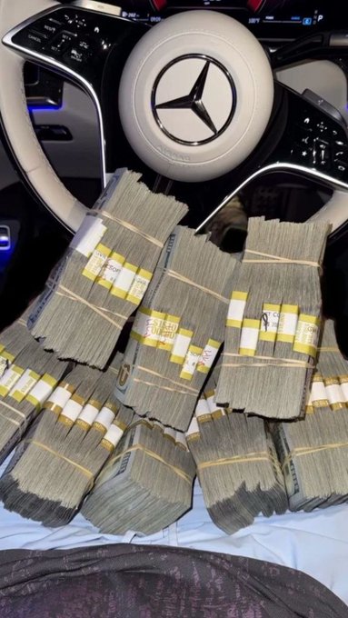 Stacks of bundled US currency piled on the dashboard of a Mercedes-Benz vehicle, identifiable by the steering wheel logo.