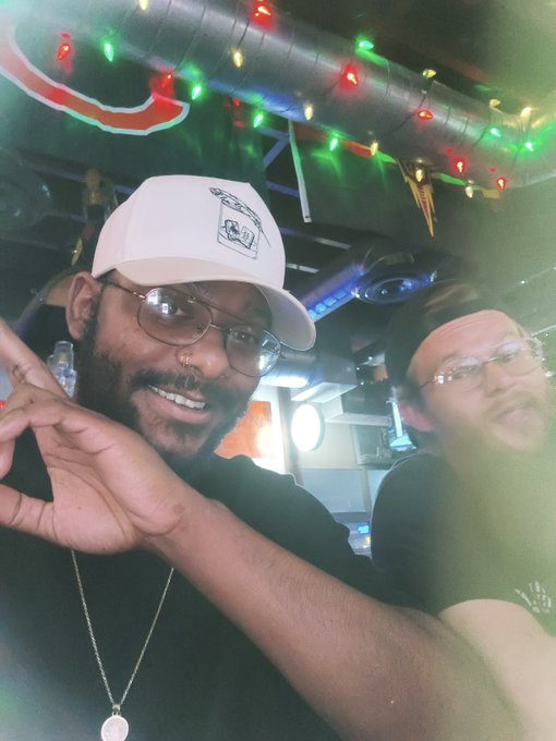A selfie of two smiling people in a dimly lit venue with colorful string lights on the ceiling, the foreground person wearing a white cap, glasses, and a black shirt, posing with a peace sign.