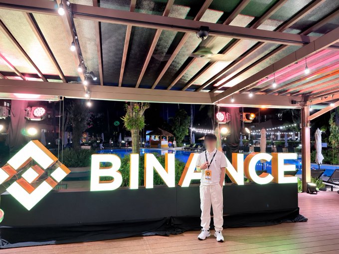 A person in white clothing stands in front of a large illuminated "BINANCE" sign with the company's logo at an outdoor nighttime venue.