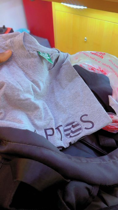 A close-up photo of folded clothing including a grey t-shirt with the 'PTS' logo printed on it, sitting on top of a pile of dark-colored clothes, with a plastic bag visible in the background.