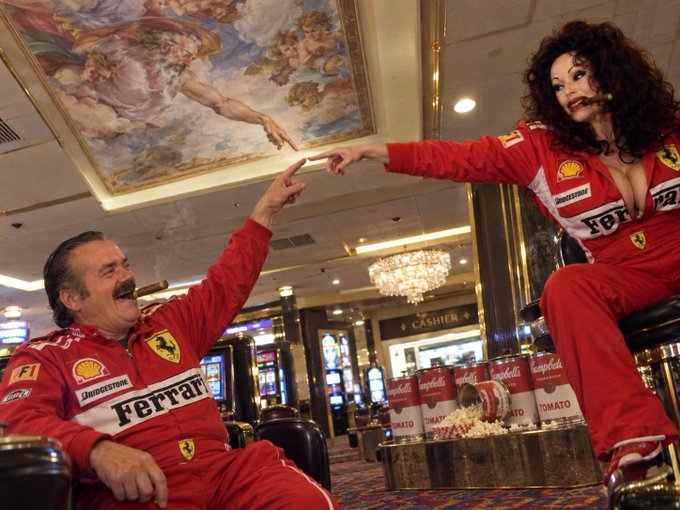 Two people wearing red Ferrari racing suits pose playfully in a casino, with the woman elevated on what appears to be a chair or stool, reaching down to touch the man's finger in a recreation of Michelangelo's 'Creation of Adam.' Campbell's soup cans sit on the table between them beneath an ornate ceiling fresco.