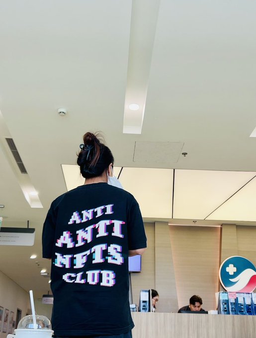 A person is seen from behind wearing a black t-shirt with the colorful text 'ANTI ANTI NFTS CLUB', standing in a brightly lit commercial space.