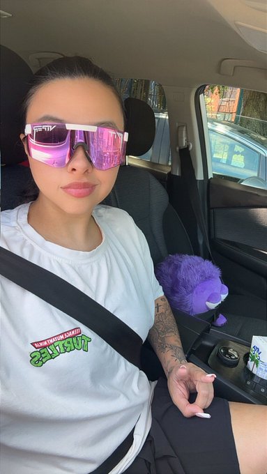 The image features a woman wearing sunglasses, sitting inside a car with her arm resting on the seat next to her. She has tattoos visible on her arms, which adds an edgy touch to her overall appearance. The photo captures a moment of self-expression through fashion and personal style.
