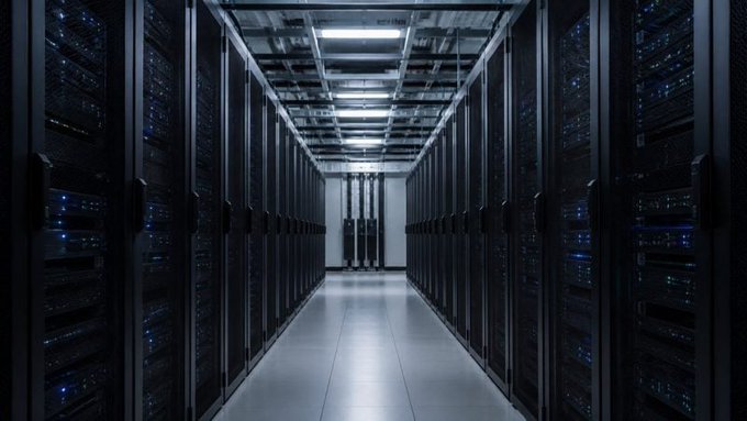 A dimly lit data center corridor lined with tall black server racks, illuminated by overhead lights and the blue glow of status LEDs.