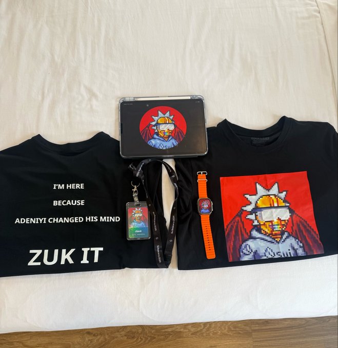 Black merchandise items are arranged on a white surface, including two t-shirts (one with 'I'M HERE BECAUSE ADENIYI CHANGED HIS MIND ZUK IT' and another with pixel art), an ID badge, an orange-strapped smartwatch, and a laptop displaying matching pixel art.