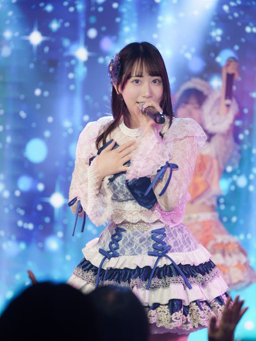 A performer on stage wearing a white and blue lace dress, holding a microphone with a sparkling blue starry background.