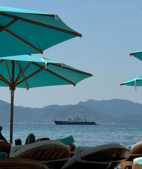 A serene beach scene featuring several teal-colored umbrellas shading wooden lounge chairs in the foreground, with people relaxing, while a boat sails across blue waters towards a mountainous coastline in the background under a clear sky.