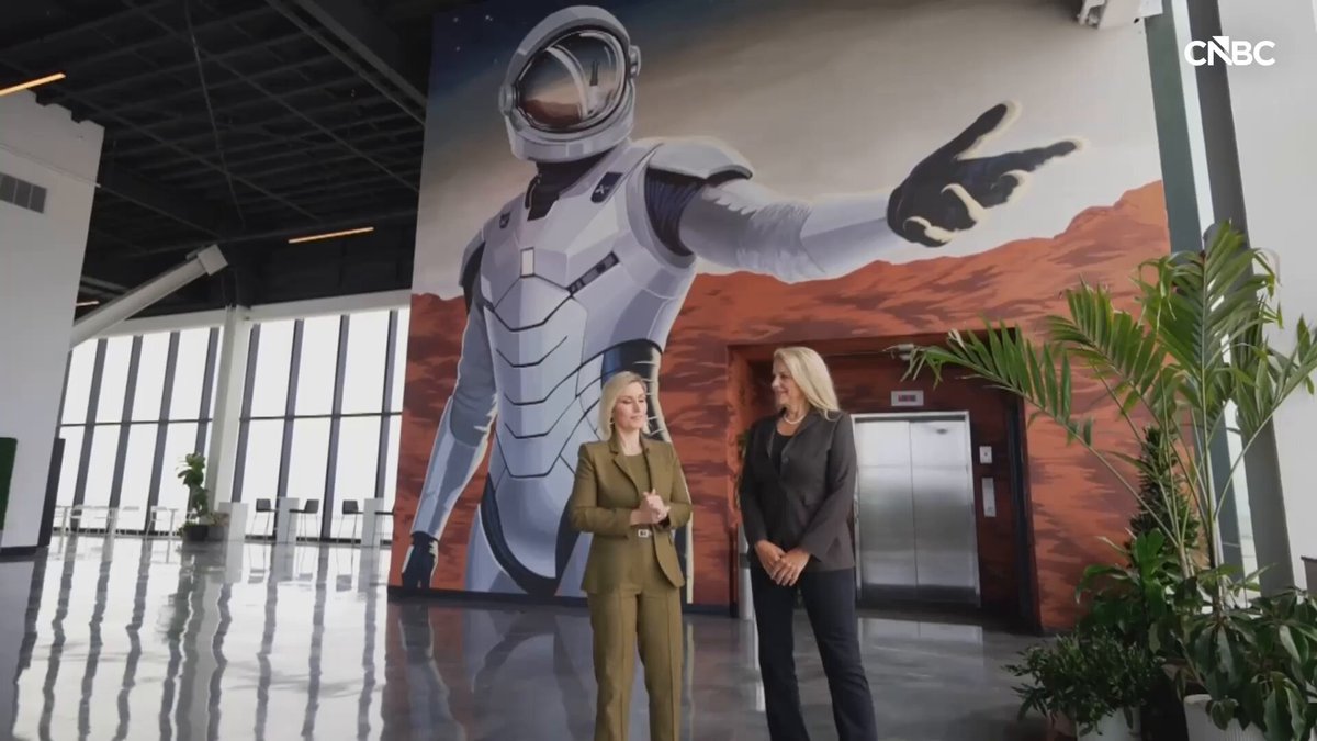 The image features two women standing next to a large mural depicting a man wearing a spacesuit. The woman on the left side of the mural has her arms crossed, while the other woman stands with her hands at her sides. They appear to be engaged in conversation or observing something interesting behind them. The mural is prominently displayed and serves as an eye-catching backdrop for their interaction.