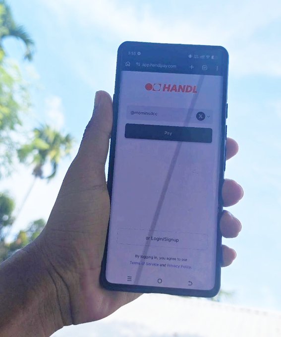 A hand holds a smartphone displaying the Handl app login page with the username '@mominsdcc' entered and a 'Pay' button visible, set against a tropical outdoor background.