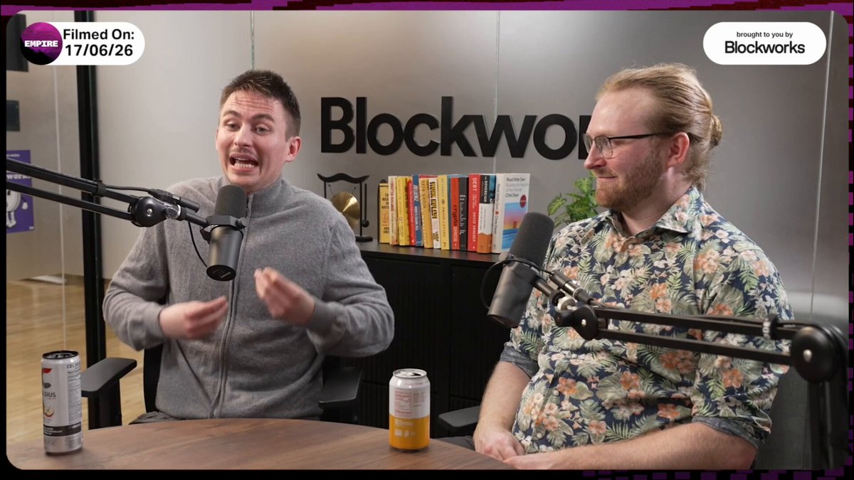 The image shows two men sitting at a table, talking into microphones during an interview or podcast recording session. The hosts are discussing the differences between Ethereum and Bitcoin as their primary competitors. One man is wearing glasses while the other has short hair. They are seated in front of a bookshelf filled with various books, which adds to the overall atmosphere of intellectual discussion.
