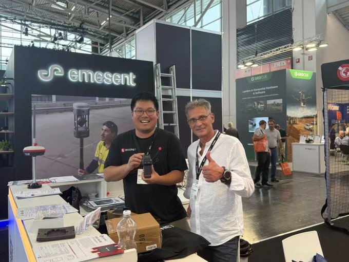Two men posing at a trade show booth for 'emesent,' with one giving a thumbs up while holding a device; a 'GEOCUE' booth is visible in the background.