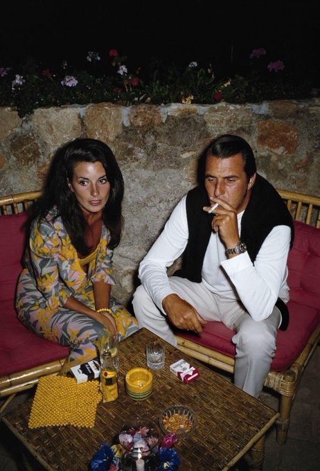 A woman and a man sit on a cushioned bench outdoors at night, with drinks and small items on a table in front of them; the man is smoking a cigarette.